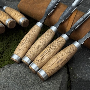 May include: A set of wood carving chisels with light brown wooden handles and silver metal collars. The blades are dark metal and curved. The chisels are arranged on a stone surface, suggesting a workshop or craft setting.