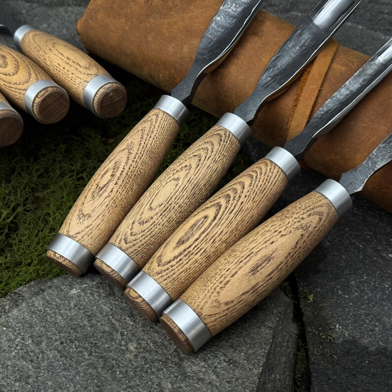May include: A set of wood carving chisels with light brown wooden handles and silver metal collars. The blades are dark metal and curved. The chisels are arranged on a stone surface, suggesting a workshop or craft setting.
