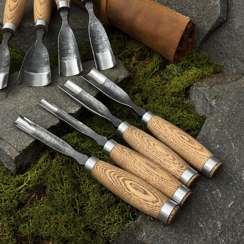 May include: A set of wood carving chisels with wooden handles and metal blades. The chisels are arranged on a stone surface with green moss, and a leather tool roll is visible in the background. The tools are designed for woodworking.