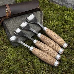 May include: A set of four metal gouges with curved, scoop-shaped blades and light brown wood grain handles with silver metal ferrules. The tools are arranged in a row, with a dark brown leather roll case visible in the upper left corner.