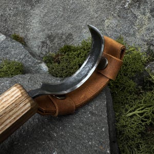 Puede incluir: Una hoja de metal curvada con un mango de madera, en una funda de cuero marrón. La hoja tiene un borde afilado y una forma curvada. La funda se asegura con un broche metálico. El fondo presenta rocas y musgo verde.