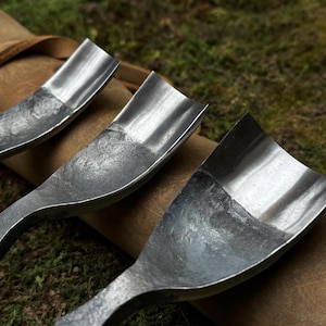 May include: Three metal carving tools with curved blades, resting on a brown leather roll. The tools have a worn, rustic look and are made of a dark metal.