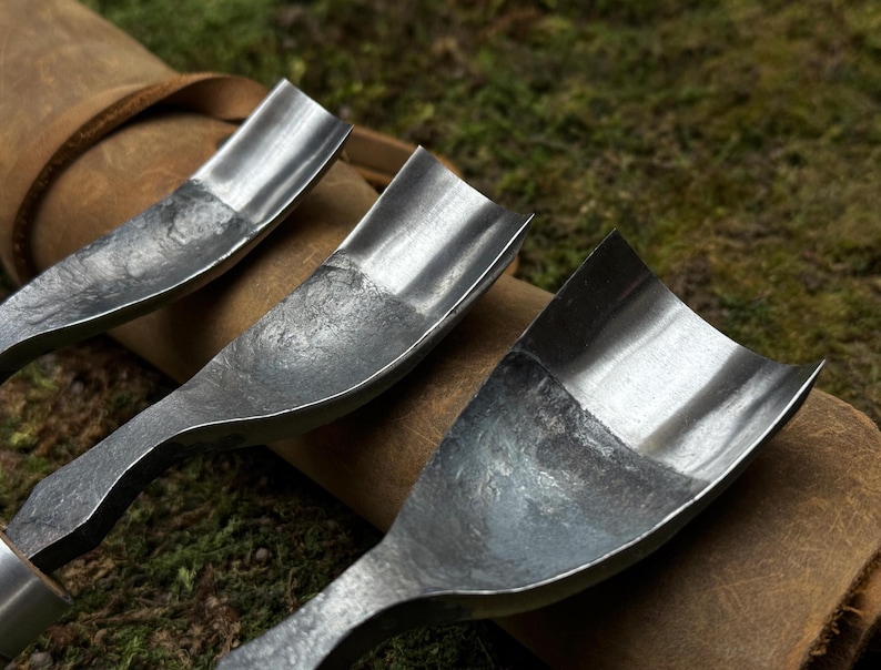 May include: Three metal carving tools with curved blades, resting on a brown leather roll. The tools have a worn, rustic look and are made of a dark metal.