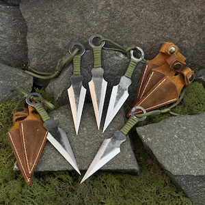 May include: Set of five silver throwing knives with green wrapped handles and leather sheaths. The knives have a sharp, triangular blade design and are displayed on a stone surface.