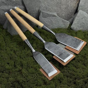 Puede incluir: Tres formones forjados a mano con mangos de madera y hojas de metal. Las hojas descansan sobre pequeñas almohadillas cuadradas de cuero. Los mangos son de madera clara con virolas de metal. Los formones están dispuestos sobre un lecho de musgo verde.