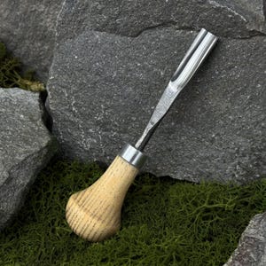 May include: A woodworking chisel with a curved blade and a light-colored wooden handle. The metal blade is silver and the handle is attached with a silver metal band. The chisel is resting on a bed of green moss and gray rocks.