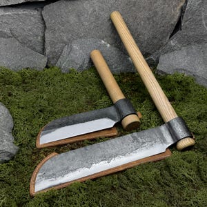 May include: Two hand-forged iron tools with light-coloured wooden handles, each featuring a curved blade edge and a protective tan leather sheath. The tools have a dark, textured metal finish and are positioned at an angle on a green surface.