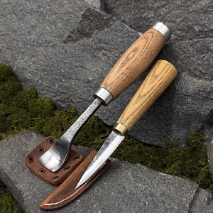 May include: Two handcrafted wood-handled tools with metal blades and leather sheaths. One tool has a curved blade, the other a straight blade. The handles are made of light-colored wood with metal accents. The tools rest on a gray stone surface.