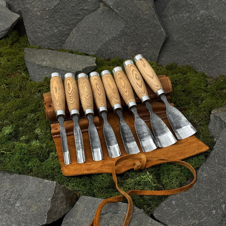 May include: A set of eight wood carving chisels with wooden handles and metal blades, displayed in a brown leather tool roll. The chisels are arranged on a bed of green moss, with large gray rocks in the background.
