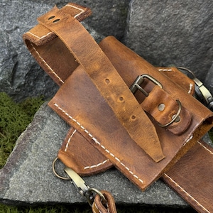 May include: Close-up of a brown leather sheath with a belt loop and strap. The sheath has a buckle closure and is stitched with white thread. The leather has a distressed look.