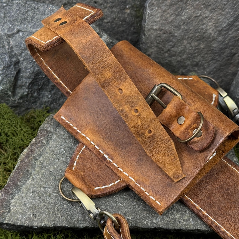 May include: Close-up of a brown leather sheath with a belt loop and strap. The sheath has a buckle closure and is stitched with white thread. The leather has a distressed look.