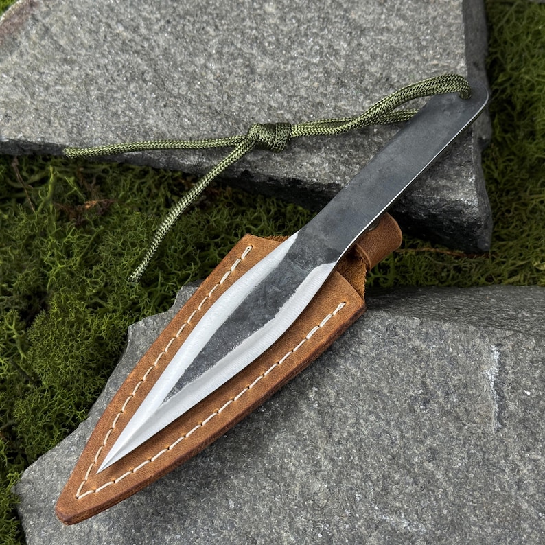 May include: A hand-forged, spear-point knife with a dark, textured blade, resting in a brown leather sheath. The sheath is stitched with white thread. A green cord is attached to the knife. The knife is displayed on a gray stone surface.