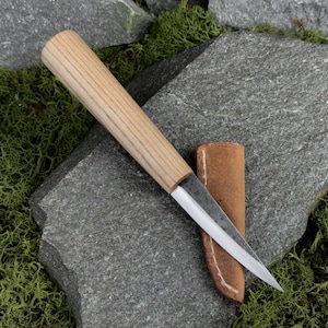 Può includere: Un coltello artigianale con un manico in legno chiaro e una lama argentata, appoggiato su una superficie di pietra grigia. Il coltello è accompagnato da un piccolo fodero in pelle marrone. Il manico è in legno chiaro.