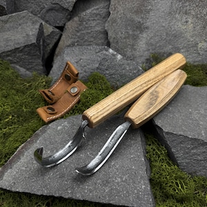 Könnte beinhalten: Zwei Holzschnitzmesser mit gebogenen Klingen und Holzgriffen liegen auf einer grauen Steinoberfläche. Eine kleine, hellbraune Lederscheide mit Metallschnappverschlüssen ist ebenfalls vorhanden. Werkzeuge für die Holzbearbeitung.