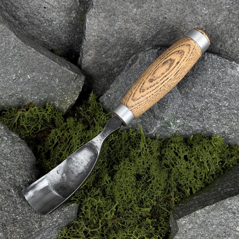May include: A wood carving gouge featuring a curved, polished steel blade and a cylindrical wooden handle with a visible grain pattern. The tool includes two silver-colored metal ferrules at the top and bottom of the handle for reinforcement.