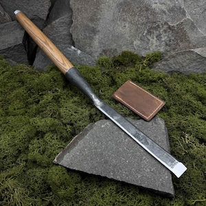 Puede incluir: Un cincel largo y delgado con mango de madera y hoja de metal. El cincel está apoyado en una roca gris, y una funda de cuero marrón está tumbada a su lado.