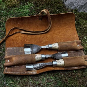 May include: A brown leather roll-up case with three wood-handled carving tools. The tools are secured in the case with leather straps.