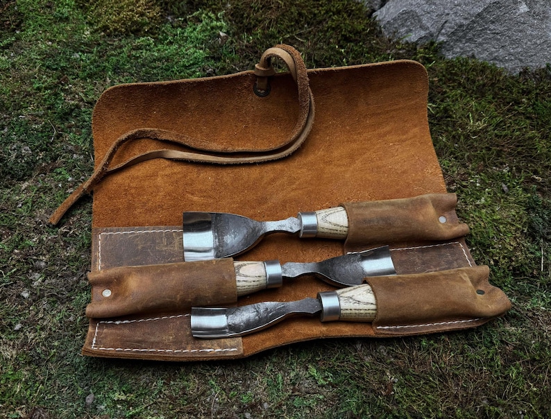 May include: A brown leather roll-up case with three wood-handled carving tools. The tools are secured in the case with leather straps.