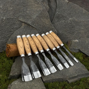 May include: A set of eight wood carving chisels with light brown wooden handles and silver metal accents. The blades are silver and angled. A brown leather tool roll is visible in the background. The chisels are arranged on a stone surface.