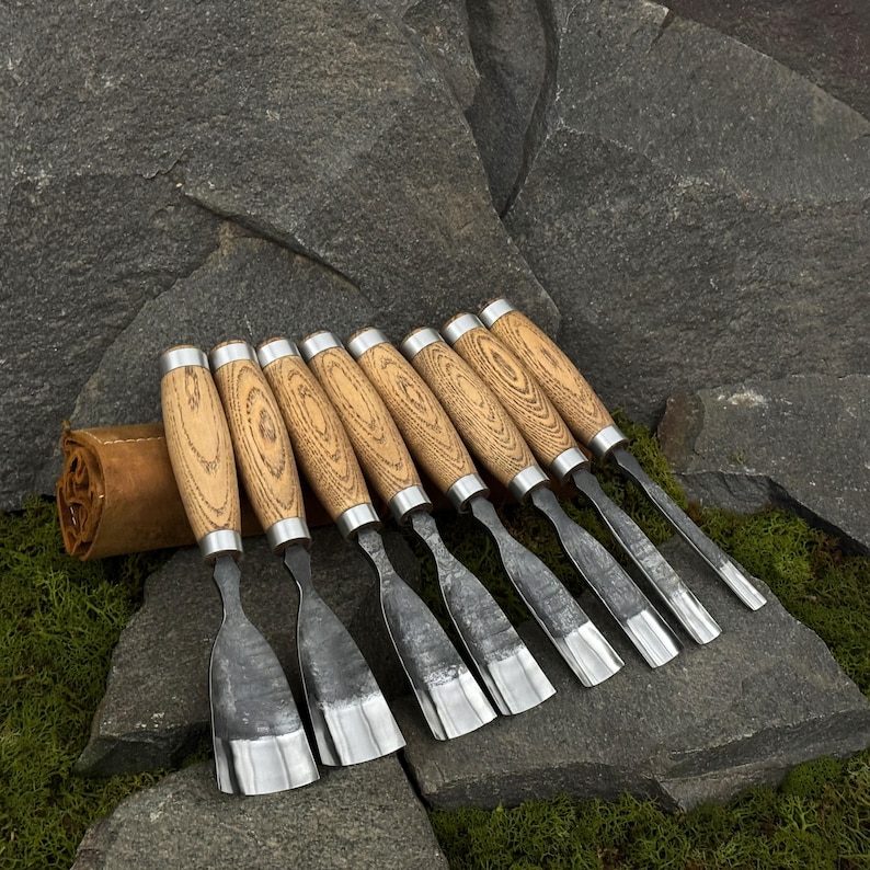 May include: A set of eight wood carving chisels with light brown wooden handles and silver metal accents. The blades are silver and angled. A brown leather tool roll is visible in the background. The chisels are arranged on a stone surface.