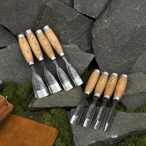 May include: A collection of wood carving chisels with light brown wooden handles and silver metal blades. The chisels are arranged on a bed of dark gray rocks and green moss, showcasing their craftsmanship.