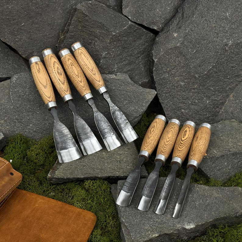 May include: A collection of wood carving chisels with light brown wooden handles and silver metal blades. The chisels are arranged on a bed of dark gray rocks and green moss, showcasing their craftsmanship.