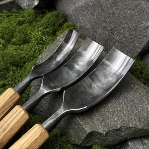 May include: Three metal gouges with curved, dark grey steel blades and light brown wooden handles. The blades feature a textured, hammered finish and are arranged side by side, showing varying widths of the curved cutting edges.