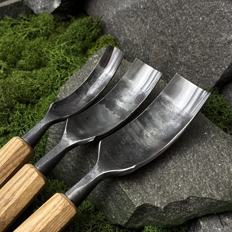 May include: Three metal gouges with curved, dark grey steel blades and light brown wooden handles. The blades feature a textured, hammered finish and are arranged side by side, showing varying widths of the curved cutting edges.