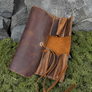 May include: A brown leather roll-up pen case with multiple compartments and a leather tie closure. The case is open, revealing the interior pockets and the natural grain of the leather. The background features rocks and green moss.