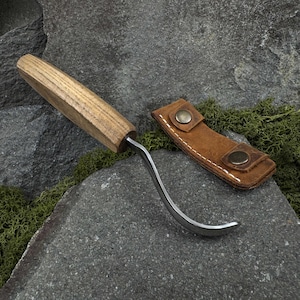 May include: A curved carving knife with a wooden handle and a metal blade, accompanied by a brown leather sheath. The knife has a distinctive hook shape, ideal for detailed woodworking. The sheath has two metal snap closures.
