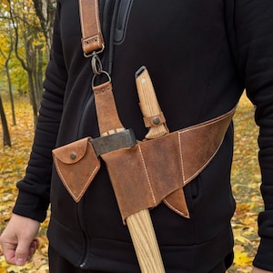 May include: A brown leather axe and knife sheath with a shoulder strap. The sheath holds a small axe and a knife with a wooden handle. The leather is stitched with white thread and has a small pouch with a snap closure.