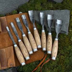 May include: A set of seven wood carving chisels with wooden handles and metal blades. The chisels are arranged on a brown leather tool roll, set against a backdrop of green moss and stone.