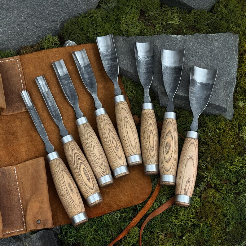 May include: A set of seven wood carving chisels with wooden handles and metal blades. The chisels are arranged on a brown leather tool roll, set against a backdrop of green moss and stone.