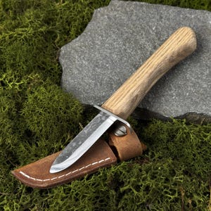 May include: A small, stainless steel knife with a light-colored wooden handle and a brown leather sheath. The knife is resting on a bed of green moss and a gray stone.
