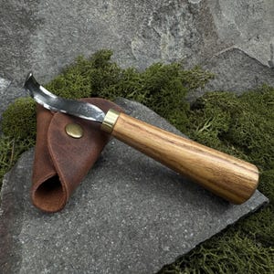 May include: A curved wood carving knife with a brown wooden handle and a brass ferrule. The knife is resting in a brown leather sheath.