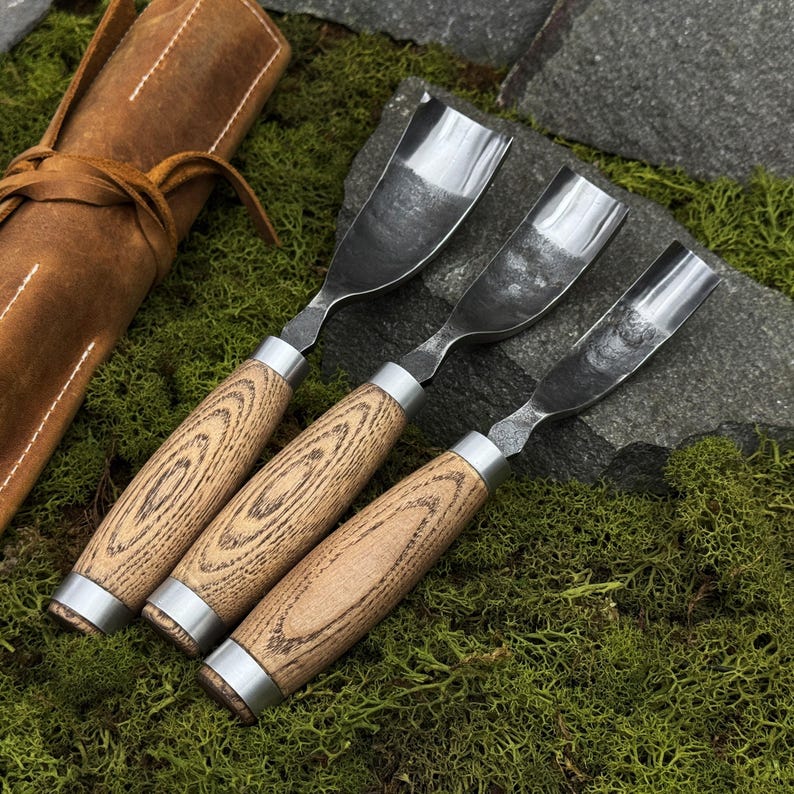 May include: Three wood carving gouges with curved metal blades and light brown wooden handles featuring a distinct grain pattern. Each tool has a silver metal ferrule connecting the blade to the handle. A brown leather tool roll is visible nearby.