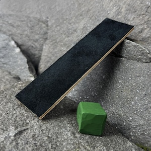 Puede incluir: Una correa de cuero negro con base de madera está en ángulo en el marco. Un pequeño objeto verde en forma de cubo se encuentra cerca de la correa. Los objetos están colocados sobre una superficie gris texturizada.