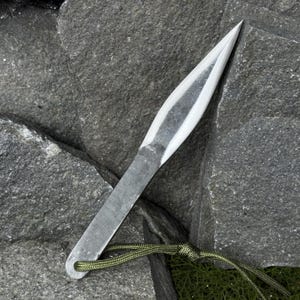 May include: A hand-forged, silver-colored throwing knife with a sharp, pointed blade. The knife has a flat, rectangular handle with a small hole at the end, and is attached to a green cord. The knife is resting on a dark gray stone surface.