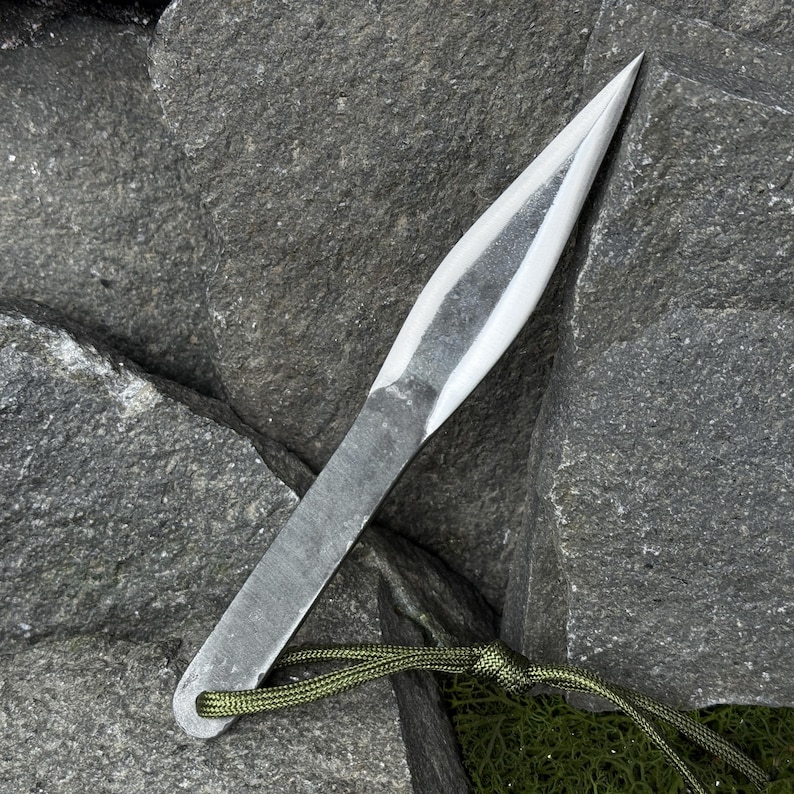 May include: A hand-forged, silver-colored throwing knife with a sharp, pointed blade. The knife has a flat, rectangular handle with a small hole at the end, and is attached to a green cord. The knife is resting on a dark gray stone surface.