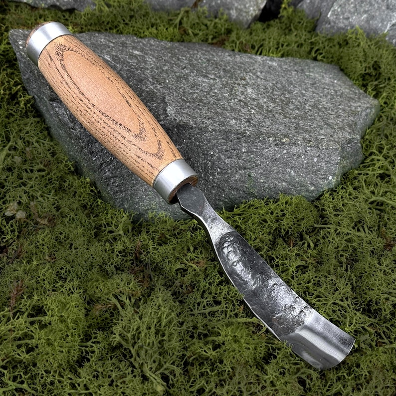 May include: A hand-forged hook knife featuring a curved, textured steel blade with a dark finish and a smooth, light-brown wooden handle secured by two polished silver-coloured metal ferrules at the top and bottom of the grip.