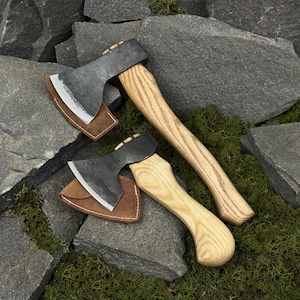 Può includere: Due piccole asce con manici in legno e lame in metallo scuro poggiano su rocce grigie. Ogni ascia ha un fodero in pelle marrone. I manici presentano un motivo a venature chiare. Le asce sono progettate per l'uso all'aperto.