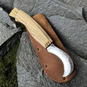 May include: A curved blade knife with a light wood handle and a brown leather sheath. The blade has a silver finish and a curved shape. The sheath has a snap closure. The knife is designed for gardening or harvesting.