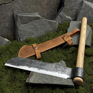 May include: A hand-forged steel brush axe with a long, light-colored wooden handle and a dark, textured blade. The tool is accompanied by a tan leather sheath featuring two brass-colored metal snap buttons and a strap for secure storage.