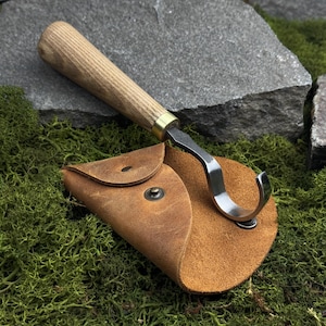 May include: A wood carving knife with a curved blade and a wooden handle, resting on a brown leather sheath. The knife has a metal blade and a brass ferrule. The sheath is closed with a snap. The knife and sheath are on a bed of green moss.