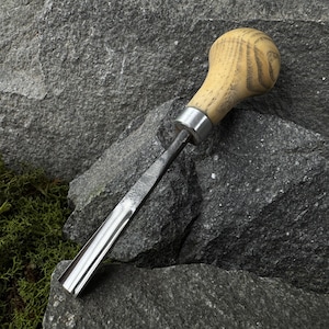May include: A woodworking chisel with a light-colored wooden handle and a silver metal blade. The handle has a rounded shape and a metal band where it meets the blade. The chisel is resting on a gray stone surface.