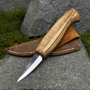 Peut inclure: Un couteau artisanal avec un manche en bois et une lame en acier inoxydable. Le couteau est présenté avec un étui en cuir marron. Le manche présente un motif de grain de bois naturel. Le couteau est conçu pour une utilisation en extérieur.
