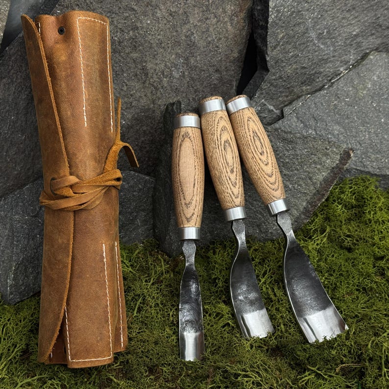 May include: A set of three wood carving tools with curved metal blades and light-colored wooden handles, accompanied by a rolled brown leather storage case featuring a tie closure and visible white stitching along the edges.
