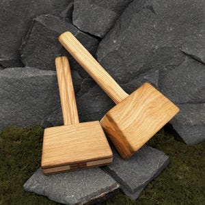 May include: Two wooden mallets with light brown handles and rectangular heads. The mallets are made of wood and have a natural finish. The mallets are resting on a dark gray stone surface with green moss.