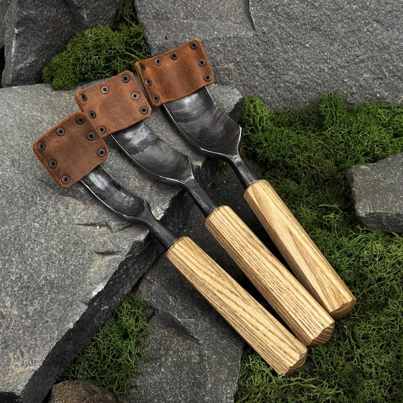 May include: Three hand-forged metal gouges with curved blades, each featuring a light-colored wooden handle and a brown leather sheath secured with metal rivets covering the sharp edge of the tool, arranged in a diagonal row on a stone surface.