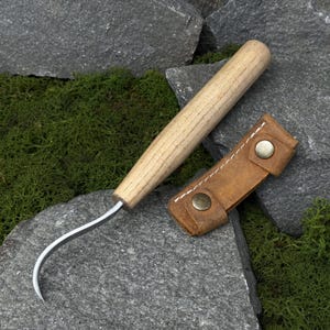 May include: A carving knife with a curved blade and a light-colored wooden handle, accompanied by a small, brown leather sheath. The knife is designed for detailed woodworking or carving projects.
